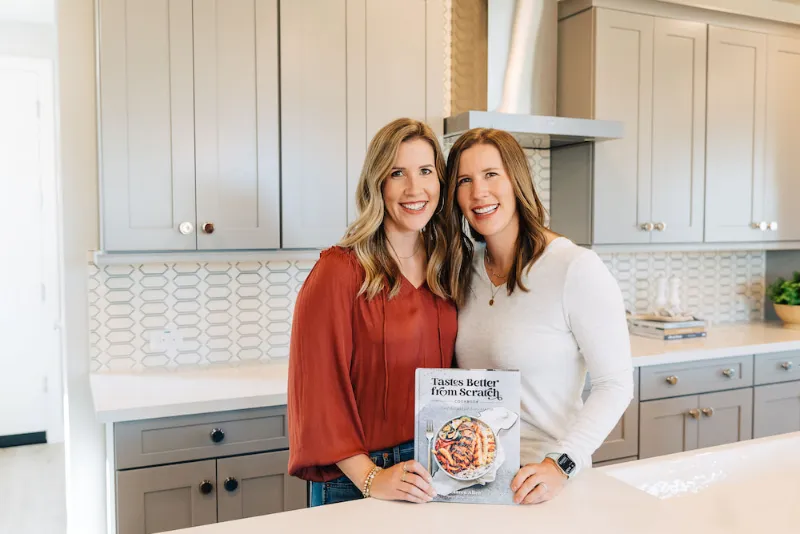 Lauren and Liz in their kitchen holding their cookbook.